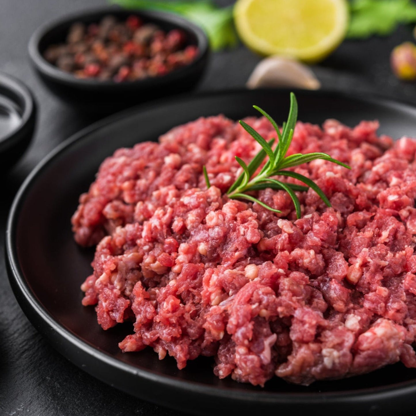 A plate of raw ground beef with a sprig of rosemary on top, surrounded by ingredients for cooking such as spices and lemon.