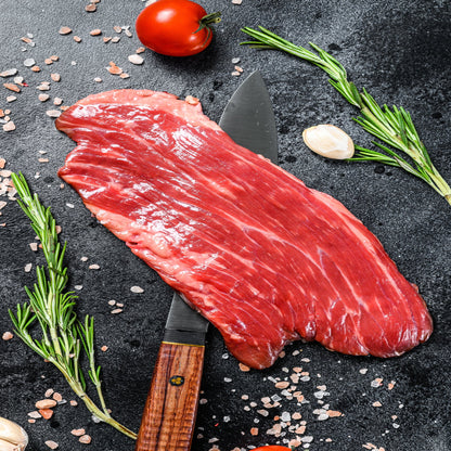 A raw flank steak on a kitchen counter surrounded by various seasonings, a tomato, and a sprig of rosemary.