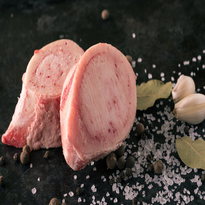 Raw Soup Bones placed on a dark surface with salt, pepper, garlic cloves, and a bay leaf scattered around.