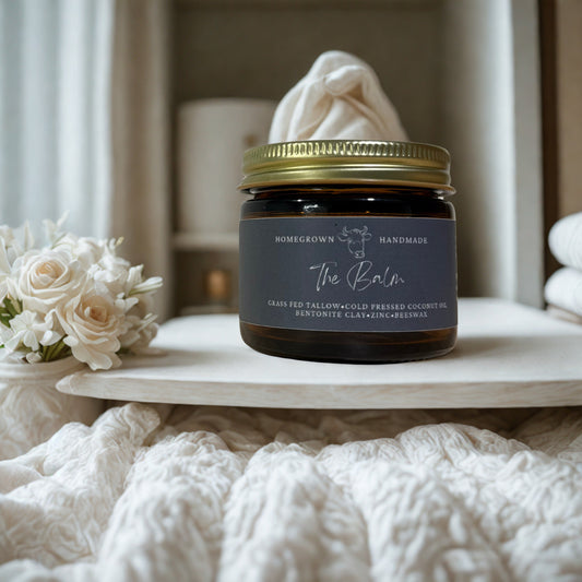 Jar of 'The Balm' with a gold lid on a white surface, surrounded by white flowers and fabric.