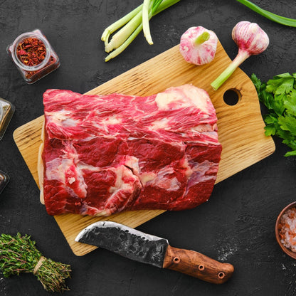 A raw chuck roast on a wooden cutting board, surrounded by various cooking ingredients such as garlic, onions, and spices, with a kitchen knife in the foreground.