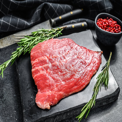 A raw flat iron steak on a stone surface with rosemary sprigs and a small bowl of seasonings beside it.